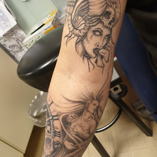 Zwart-wit tattoos van een gehoornde vrouw met schedel en een vrouw met klok op de arm.