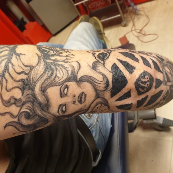 Zwart-wit tattoo van een vrouwengezicht met golvend haar en een geometrisch '666' patroon.