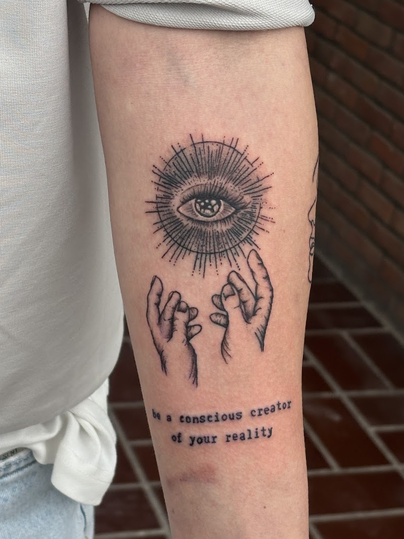 Zwart-wit tattoo van een oog met stralen, twee handen en tekst op de onderarm.