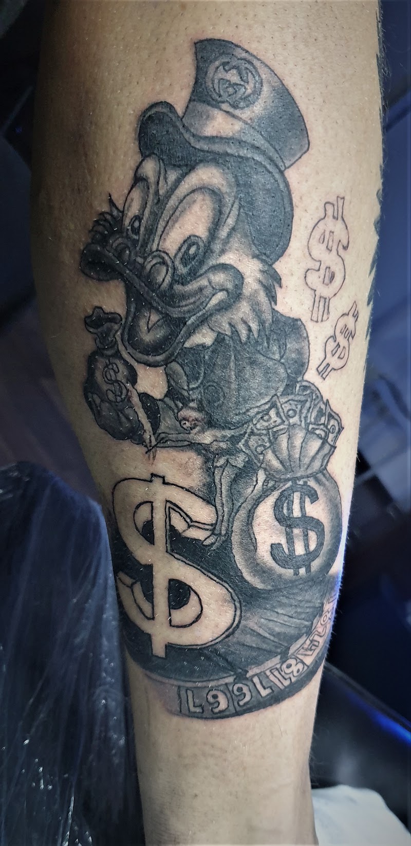 Zwart-wit tattoo van Dagobert Duck met geldzakken en dollartekens op onderbeen.