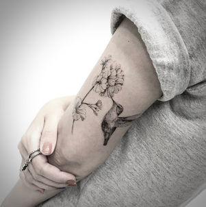 Fineline tattoo van een kolibrie bij bloemen op de bovenarm.