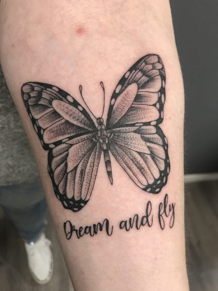 Zwart-wit vlinder tattoo met de tekst "Dream and fly" eronder.