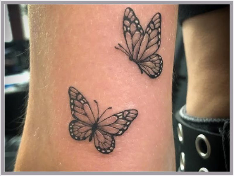Twee kleine, gedetailleerde vlinder tattoos in zwart-wit op de enkel.