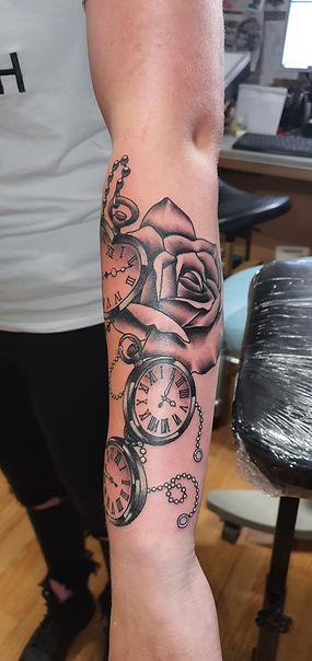 Zwart-wit tattoo van een roos en drie zakhorloges op de onderarm.