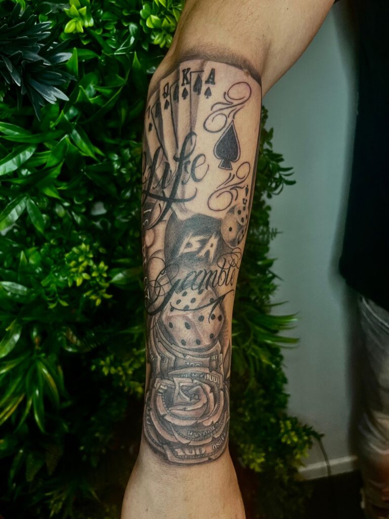 Zwart-wit gokthema tattoo met kaarten, dobbelstenen, roos en geld op onderarm.