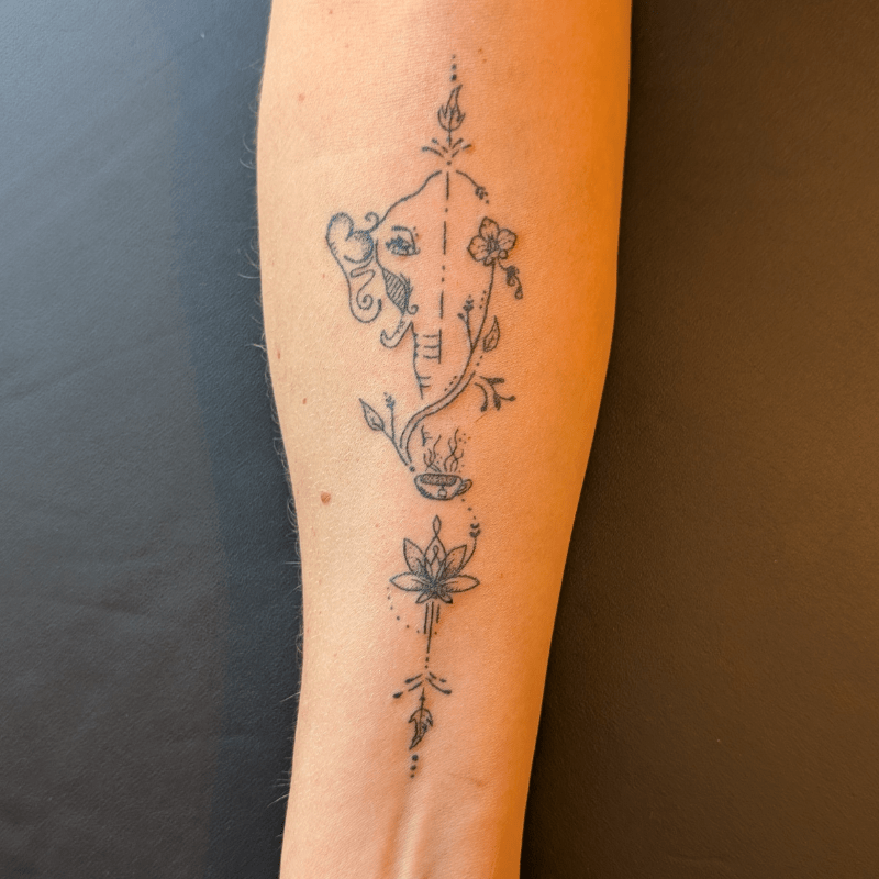 Fineline tattoo van Ganesha, lotus, bloemen en een kopje op de onderarm.