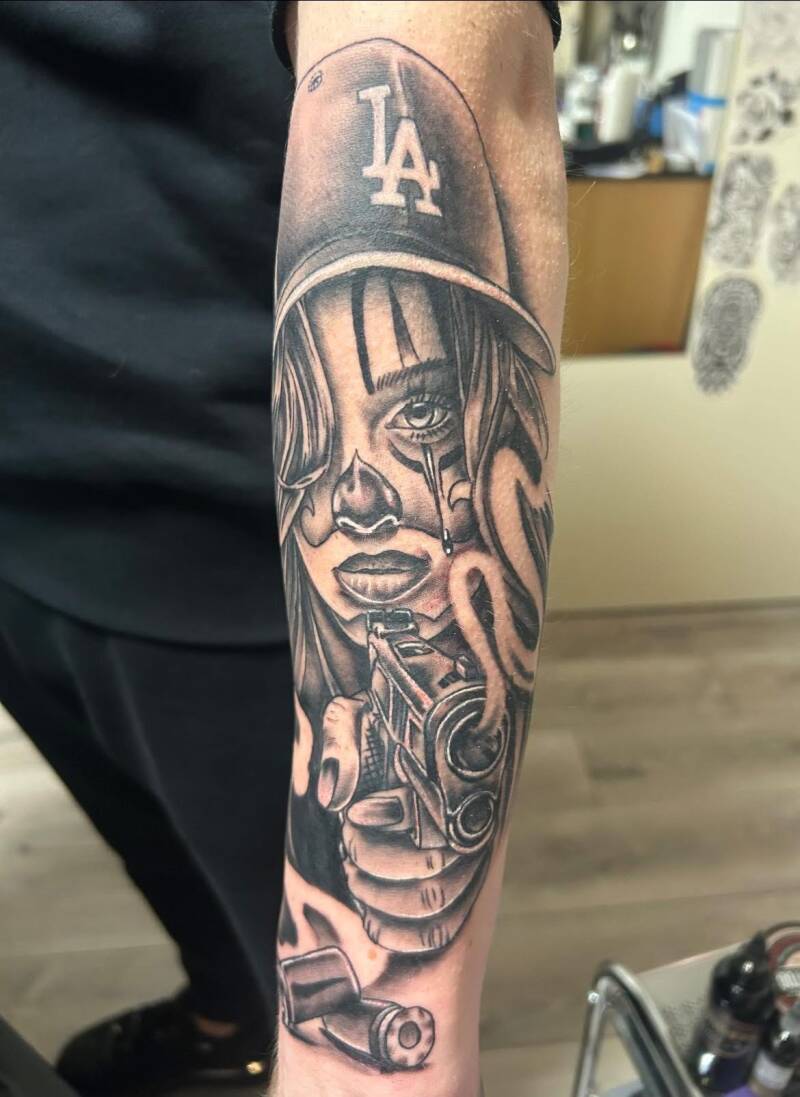 Chicano stijl tattoo van vrouw met LA pet en pistool op onderarm.