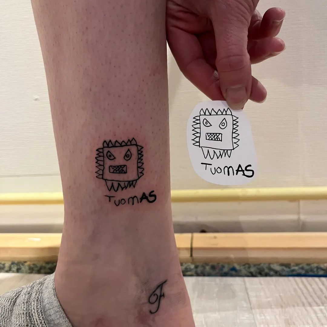 Kleine kindertekening tattoo van een monster en de naam 'Tuomas' op de enkel.