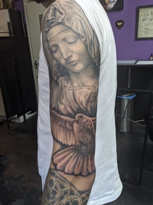 Zwart-wit tattoo van de Maagd Maria en een duif op de bovenarm.