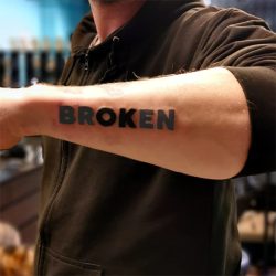Duidelijke 'BROKEN' tekst tattoo in blokletters op de onderarm.