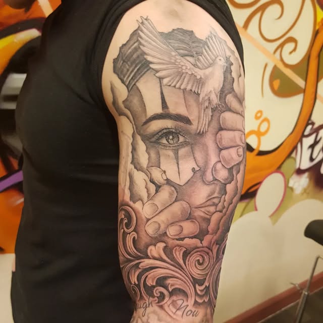 Zwart-wit tattoo van een gezicht met duif, handen en filigraan op de bovenarm.
