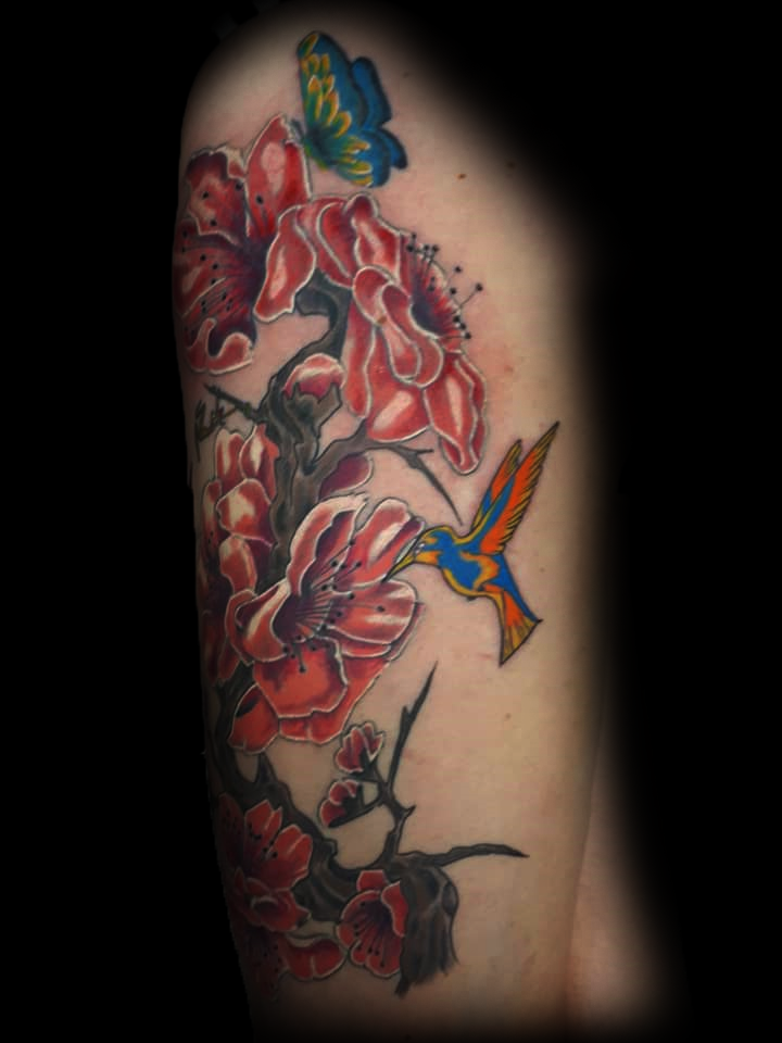 Kleurrijke tattoo van rode bloemen, een kolibrie en een vlinder op een tak.