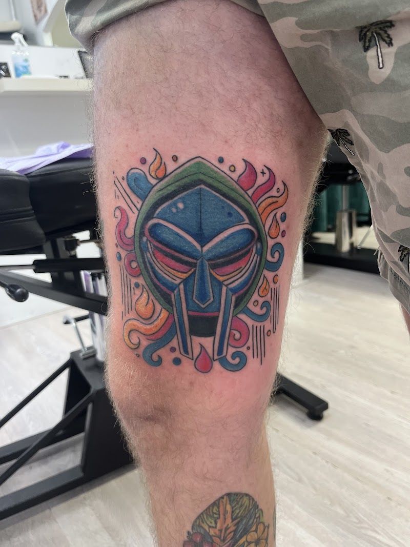 Kleurrijke illustratieve tattoo van het MF DOOM masker op het bovenbeen.