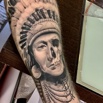 Realistische tattoo van een Indiaan met een half schedel gezicht op de onderarm.