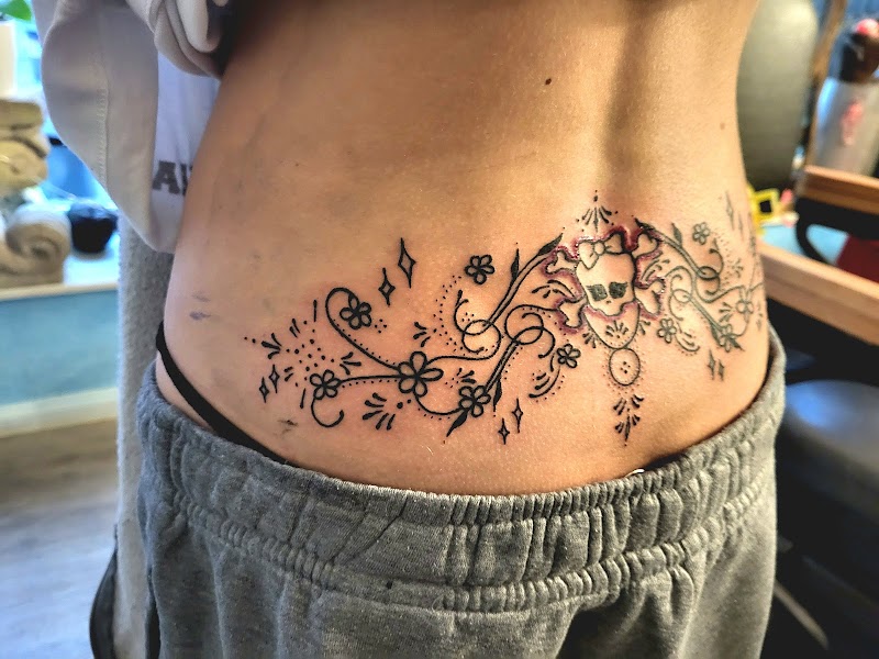 Sierlijke onderrug tattoo met schedel, bloemen en krullen in fineline stijl.