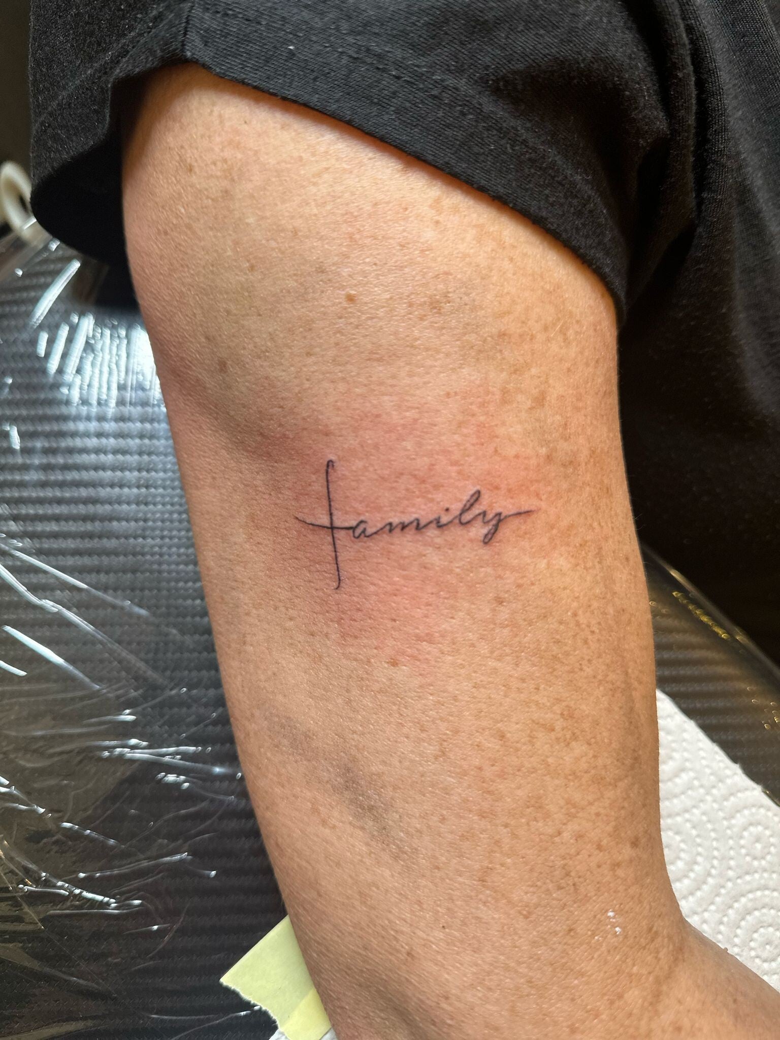 Minimalistische 'family' tekst met kruis op bovenarm.