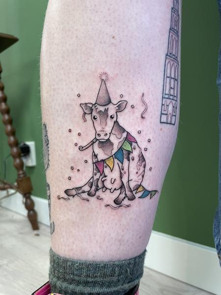 Illustratieve tattoo van een feestende koe met hoedje en slingers op onderbeen.