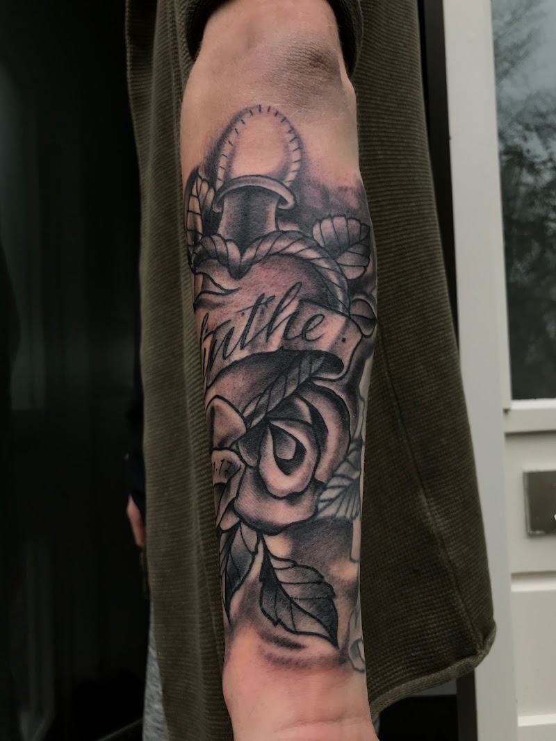 Zwart-wit tattoo van een anker, roos en spandoek met tekst op de onderarm.