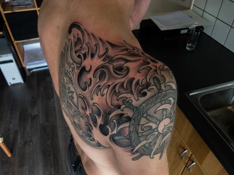 Zwart-wit tattoo van Japanse golven en een stuurwiel op de schouder.