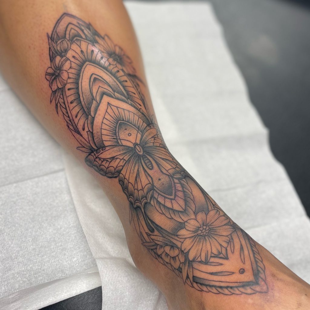 Grote ornamentale tattoo met vlinder en bloemen op onderbeen.