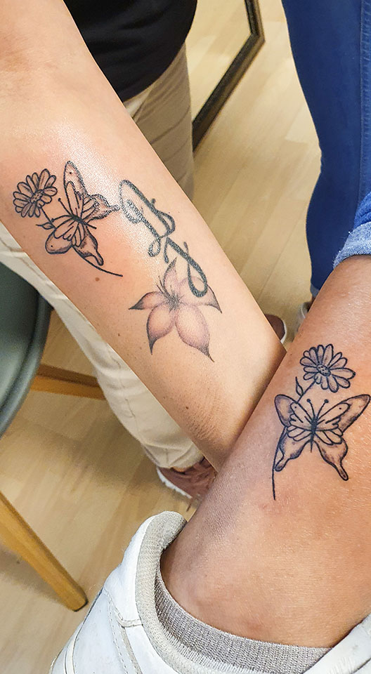 Twee onderbenen met bijpassende vlinder en bloem tattoos, plus een initiaal en lelie.