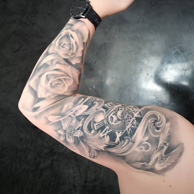 Zwart-grijze tattoo met rozen, lelies, een klok en een duif op de bovenarm.