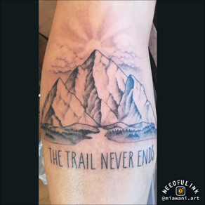Berglandschap met zon en tekst "THE TRAIL NEVER ENDS" op onderbeen.