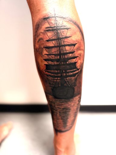 Zwart-wit tattoo van een zeilschip silhouet tegen een volle maan op het onderbeen.
