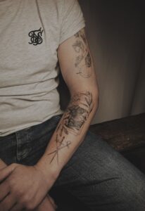 Zwart-wit tattoos op arm: landschap in kader, gekruiste pijlen, bloem.