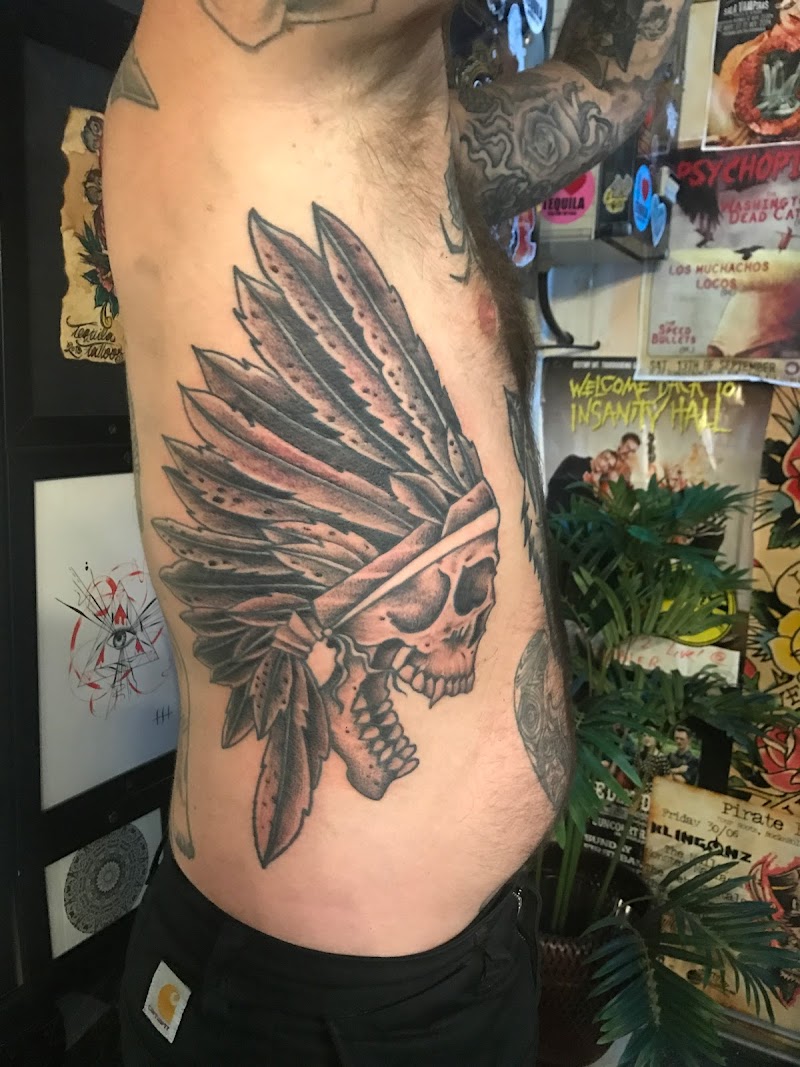 Grote zwart-wit tattoo van een schedel met een indianen verentooi op de ribben.