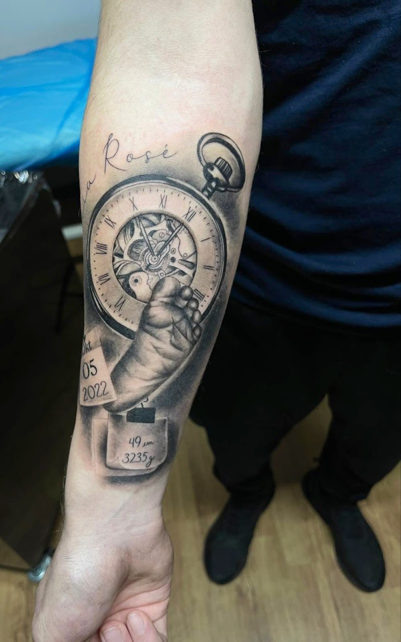 Realistische tattoo van zakhorloge, babyvoetje en geboortegegevens op onderarm.