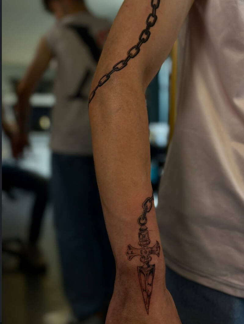 Zwart-wit tattoo van een ketting die om de arm wikkelt met een dolk/kruis hanger.