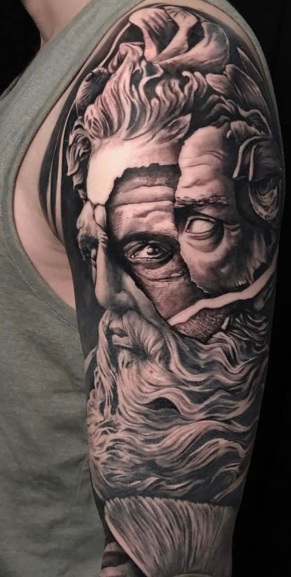 Realistische zwart-wit tattoo van een gebarsten Grieks godenhoofd op de bovenarm.