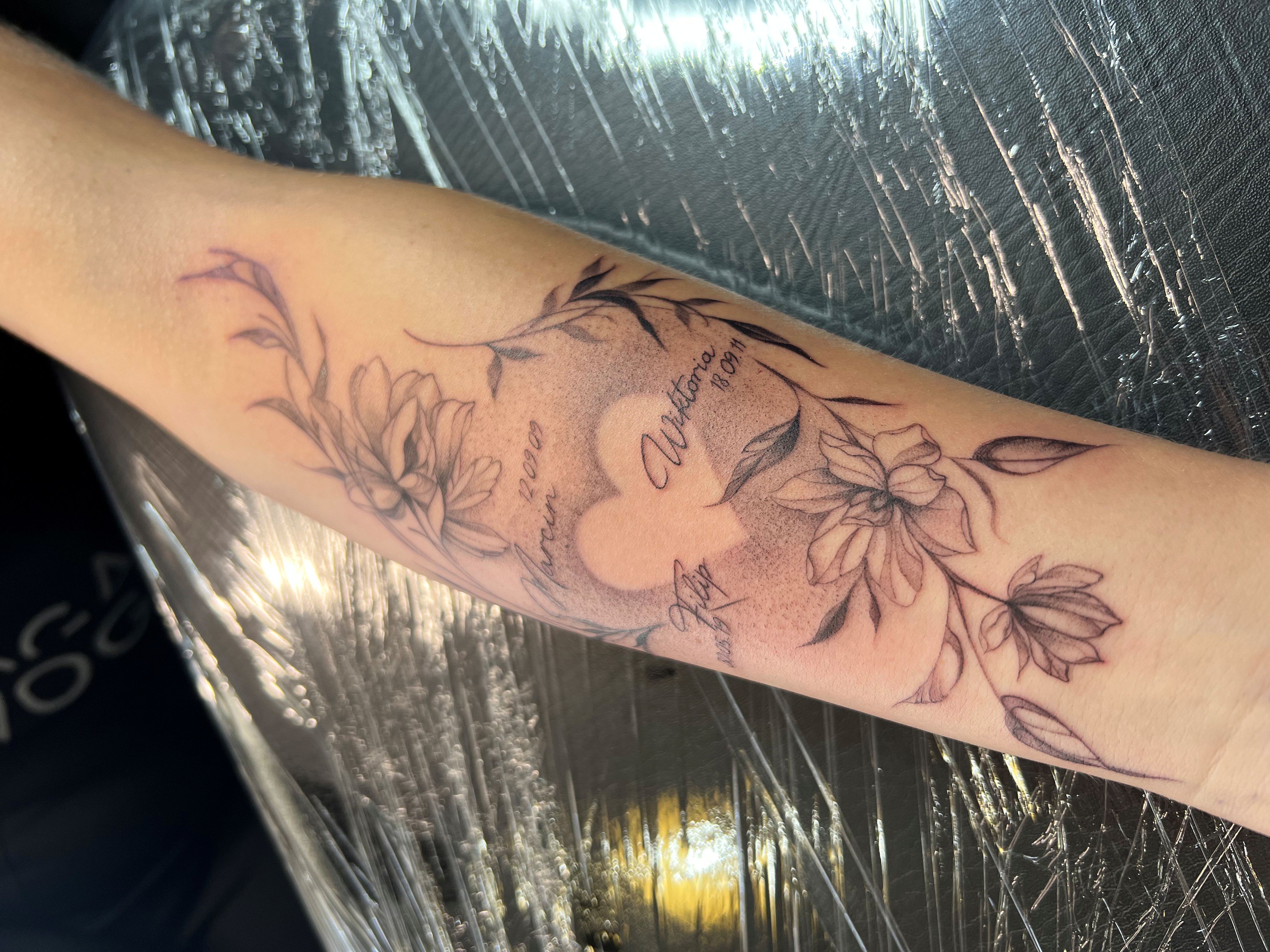 Fineline bloemen en hart met namen en datums op onderarm.