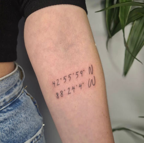Fineline tattoo van coördinaten op de achterkant van de bovenarm.
