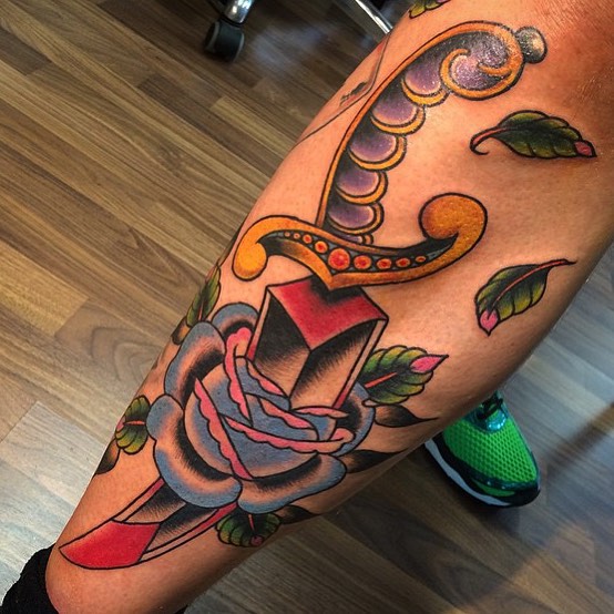 Traditionele tattoo van een dolk door een blauwe roos op het onderbeen.