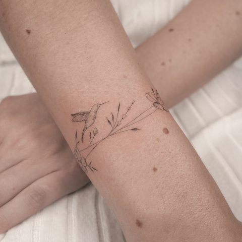Fineline tattoo van een kolibrie en bloemen die om de pols wikkelt.
