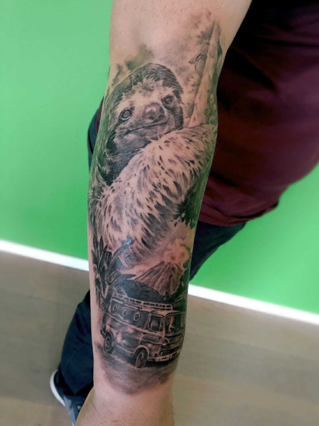 Zwart-wit tattoo van luiaard, vulkaan en VW bus op onderarm.