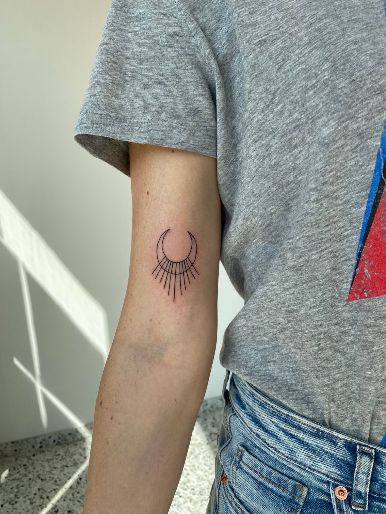 Minimalistische fineline tattoo van een halve maan met stralen op de bovenarm.
