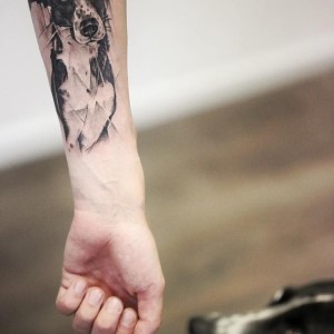 Zwart-wit tattoo van een hond met geometrische en schetsmatige elementen op de onderarm.