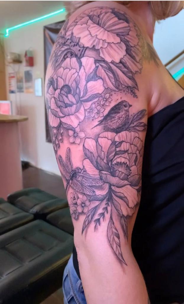 Zwart-wit tattoo van pioenrozen, een vogel en een libel op de bovenarm.