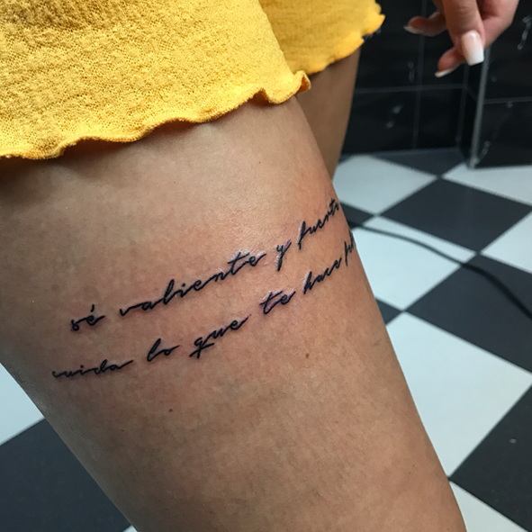 Fineline tekst tattoo op bovenbeen met Spaanse quote.