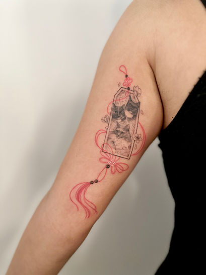 Fineline tattoo van een omamori amulet met drie kattenportretten en rode touwtjes.