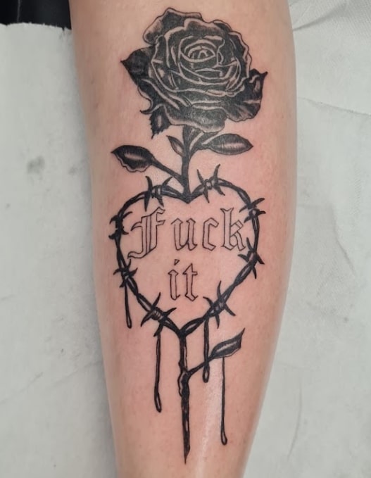 Zwart-wit tattoo van een roos in een hart van prikkeldraad met tekst 'Fuck it'.