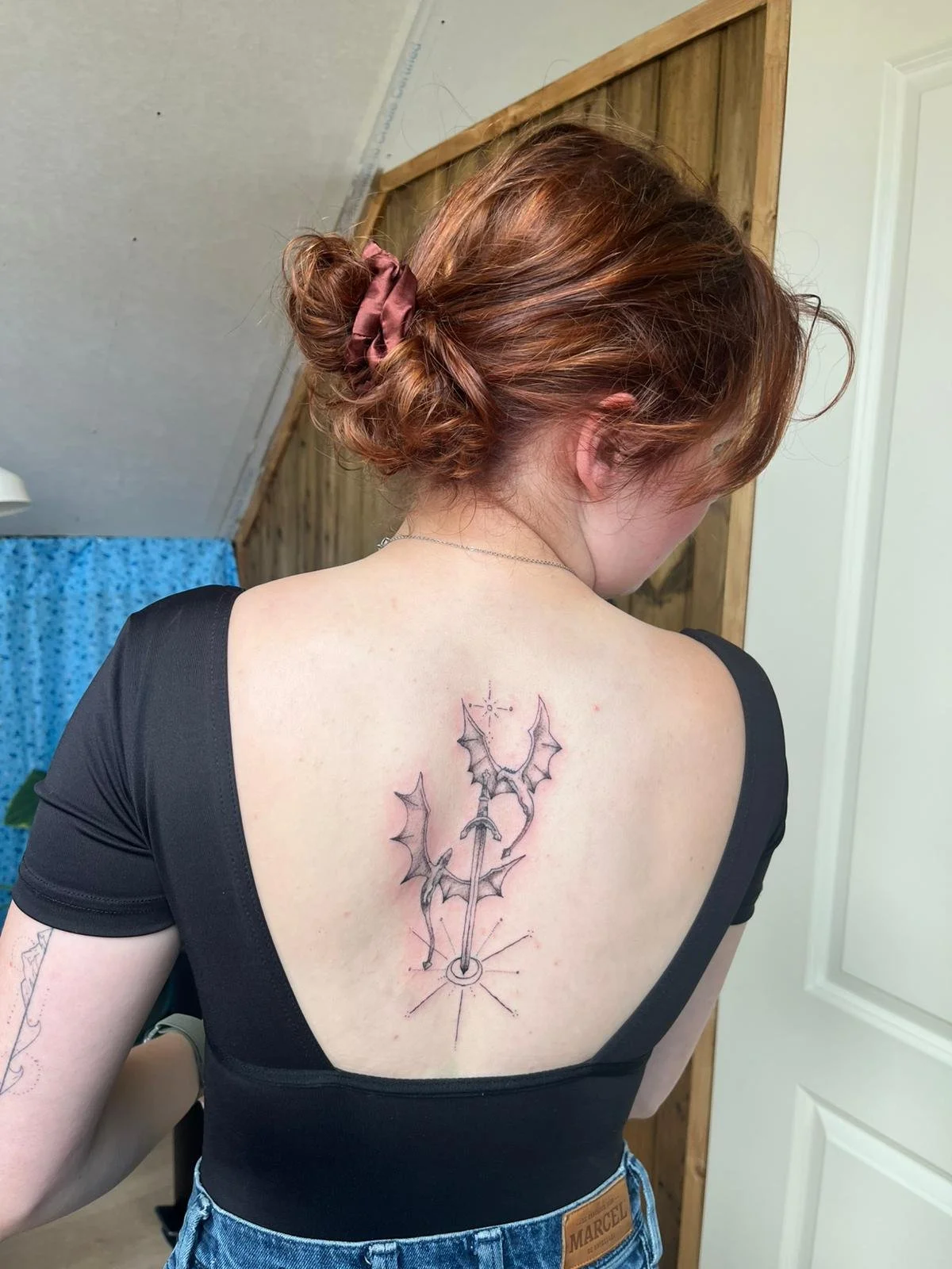 Fineline tattoo van een zwaard met drakenvleugels op de bovenrug.