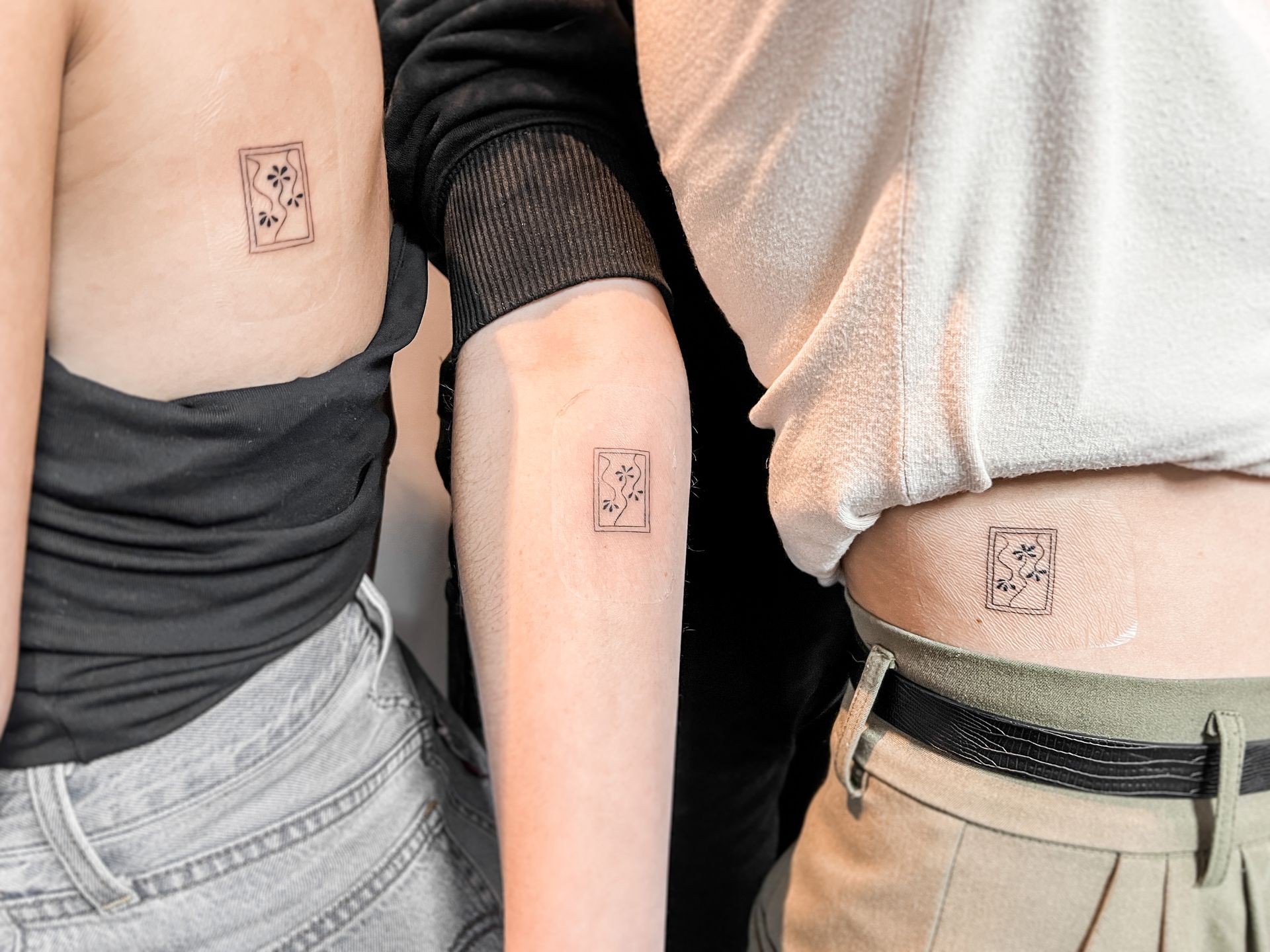 Drie identieke fineline tattoos van een bloem in een kader op ribben, arm en heup.