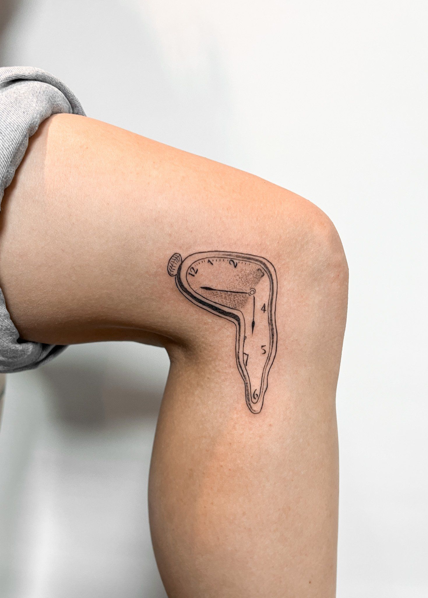 Fineline tattoo van een smeltende Dalí klok op de zijkant van de knie.