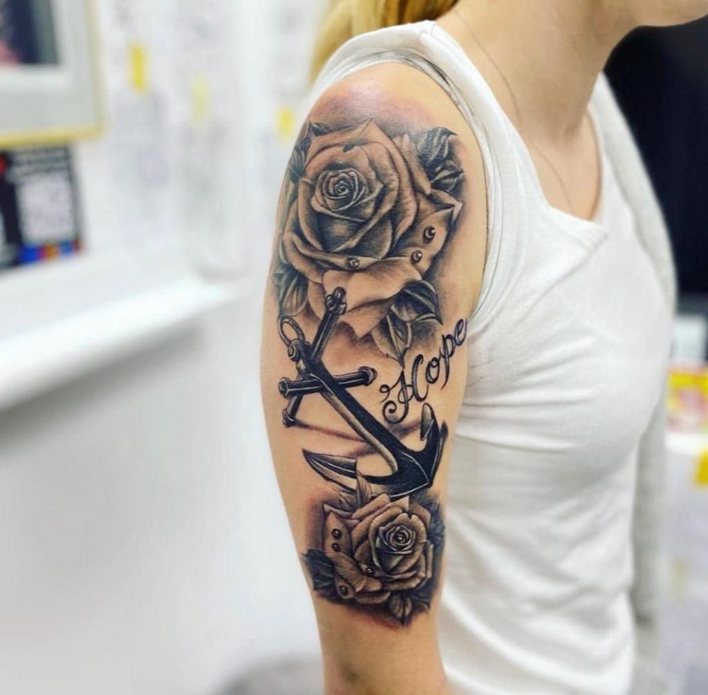 Zwart-grijze tattoo van rozen, een anker en het woord 'Hope' op de bovenarm.