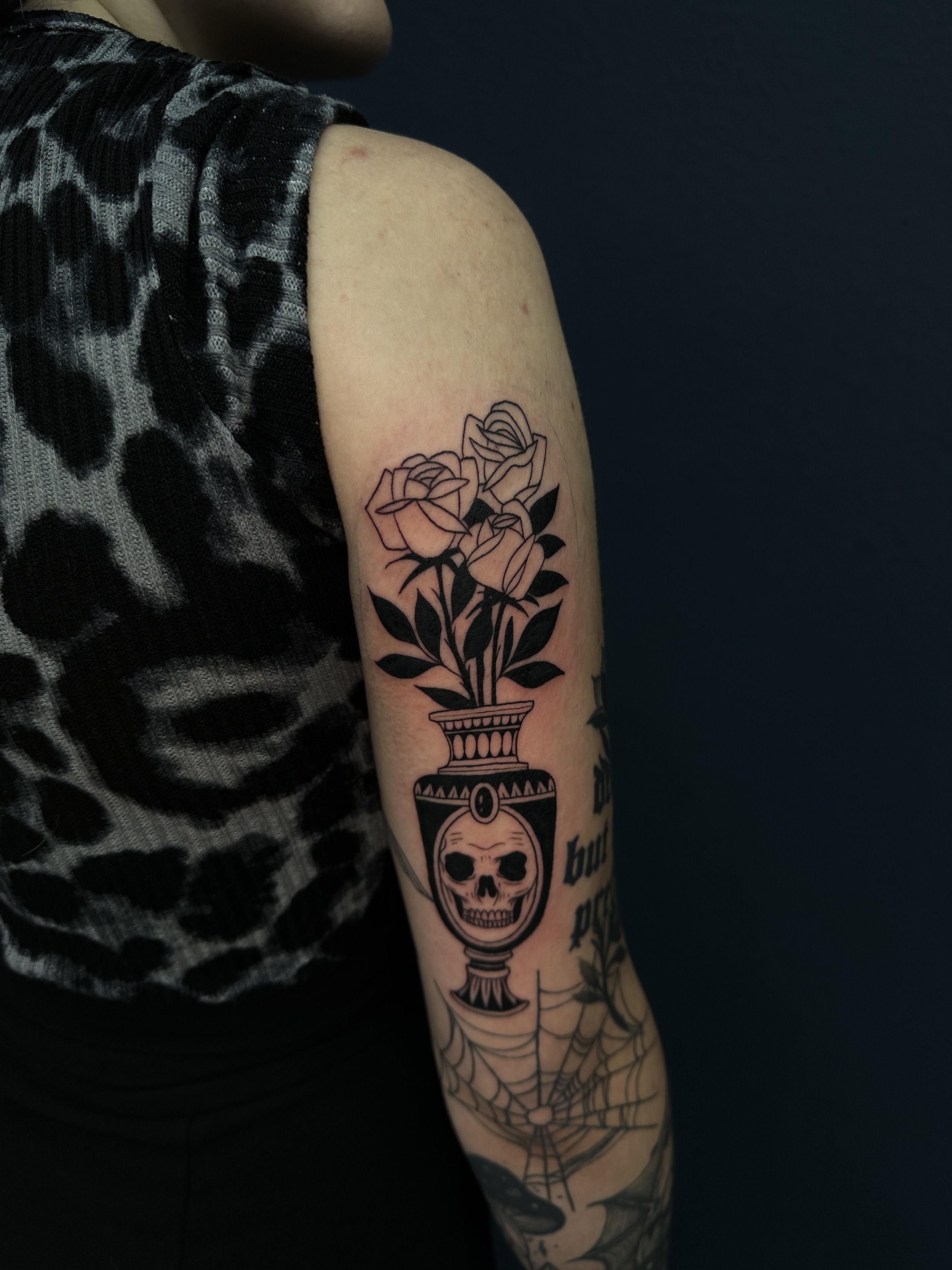 Zwart-wit tattoo van een vaas met schedel en rozen op de bovenarm.