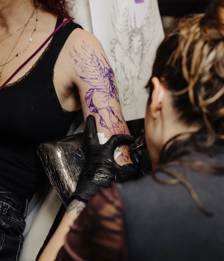 Tattoo artiest zet een schetsmatige gevleugelde tattoo op een bovenarm.